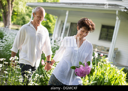 Organic farm. Summer party. A mature couple in white shirts walking holding hands through the garden. Stock Photo