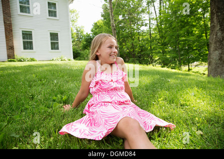 A young girl in a pink patterned sundress sitting on the grass under the trees in a farmhouse garden. Stock Photo