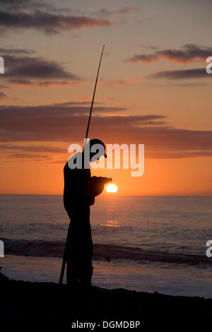 A fisher knotting a fishing line in front of the setting sun, Gillespie Beach, South Island, New Zealand Stock Photo