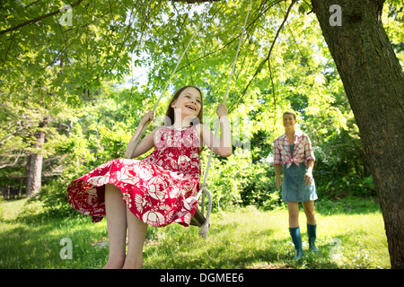 Summer. A girl in a sundress on a swing under a leafy tree. A woman standing behind her.