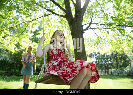 Summer. A girl in a sundress on a swing hanging from a tree branch. A woman behind her.