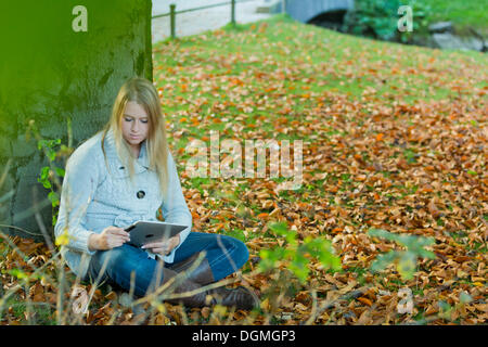 Young woman leaning against a tree in the park and looking at a tablet PC Stock Photo