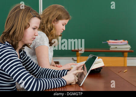 Two schoolgirls in the classroom, one is reading a book, the other is using a tablet PC, Germany Stock Photo