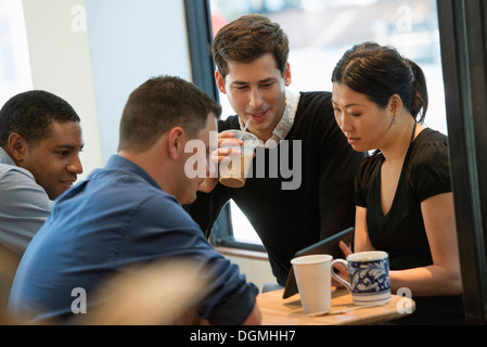 A group of people sitting around a table in a coffee shop. Looking at the screen of a digital tablet. Three men and a woman. Stock Photo