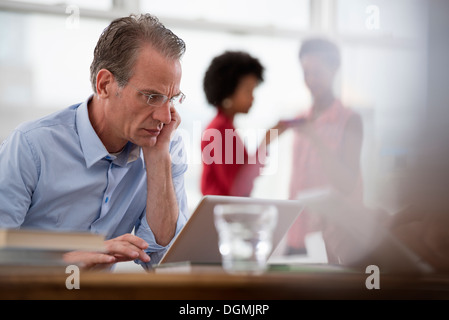 Office life. A man seated at a computer laptop leaning on one arm, and two women in the background. Stock Photo