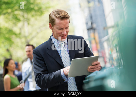 Three business people outdoors in the city. A man using a digital tablet, and a couple in the background. Stock Photo