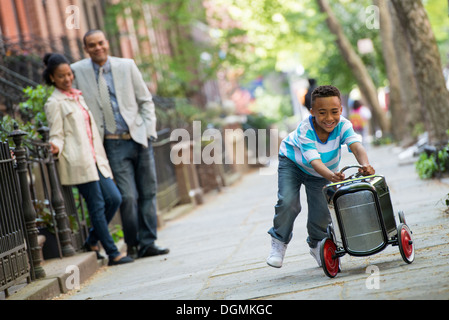 A young boy playing with a old fashioned toy car on wheels on a city street. A couple looking on.