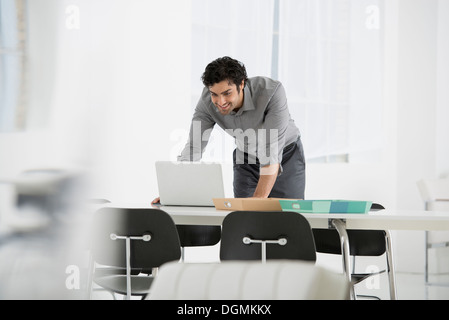Business. A man standing over a desk, leaning down to use a laptop computer. Stock Photo