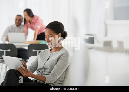 Business. A woman sitting down using a digital tablet. Two people in the background looking at a computer screen. Stock Photo