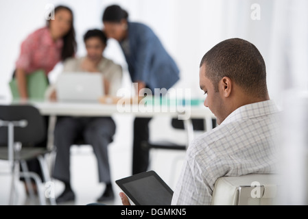 Business. A man seated in the foreground, and three people working at a laptop. Stock Photo