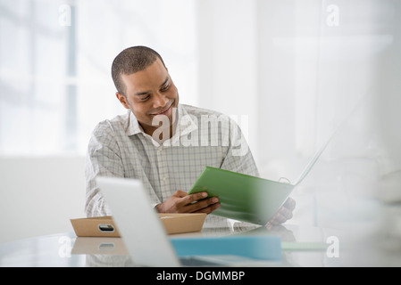 Business. A man seated at a desk opening a file of paperwork. A laptop open. Stock Photo