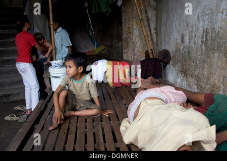 A young child laborer boy living in poverty is sitting on a wooden bed while others are sleeping in Kampong Cham, Cambodia. Stock Photo