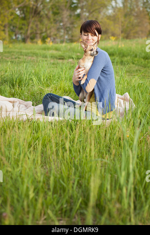 A young woman sitting in a field, on a blanket, holding a small chihuahua dog.