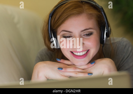 Smiling redhead lying on the couch with her laptop listening to music Stock Photo