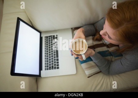 Redhead lying on the couch looking at laptop having coffee Stock Photo