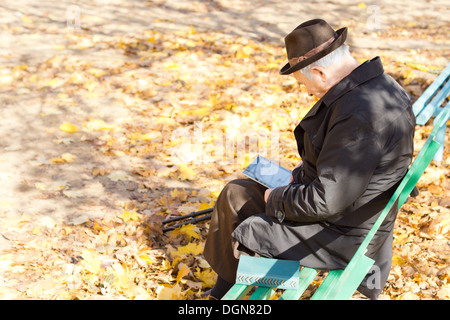 Elderly one legged handicapped man sitting reading in the park on a wooden bench on a sunny autumn day with his crutches alongside him. Stock Photo