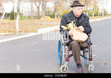 Elderly man in a wheelchair bring home the groceries from the supermarket in a brown paper bag as he sits waiting in the street Stock Photo