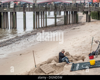 Sculptor creating sand sculpture of a man reclining on a sofa, on River Thames South Bank, London, UK for tips Stock Photo