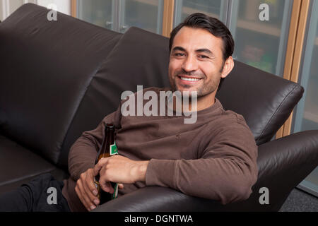 Smiling man sitting with a bottle of beer on a leather sofa Stock Photo