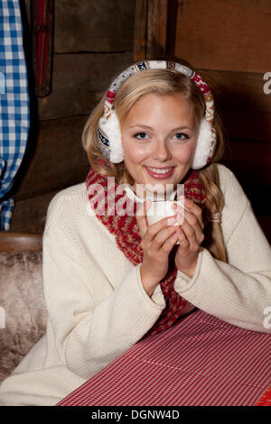 Smiling young woman wearing ear warmers and a scarf sitting at the table of a hiking hut and drinking a hot drink Stock Photo