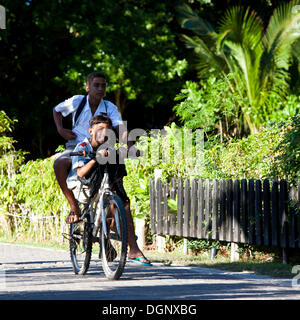 Two boys in school uniforms on a bicycle on their way to school, La Digue, Seychelles, Africa, Indian Ocean Stock Photo