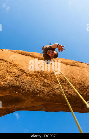Young man abseiling from a rock arch, hanging upside down, near the Spitzkoppe granite peaks, Damaraland, Namibia, Africa Stock Photo