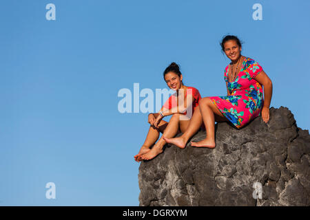 Two young women, ca. 15 and 19, sitting on a rock in front of the blue sky, Sukadana, Tukad Batang, Bali, Indonesia Stock Photo