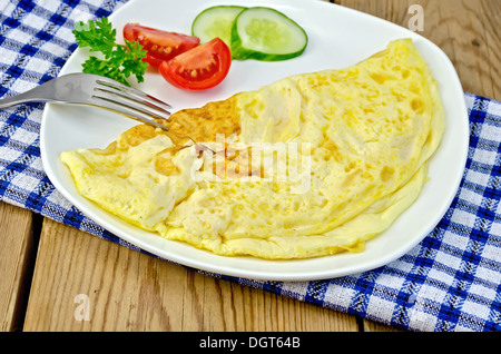 Omelette with slices of tomato, cucumber, parsley and fork on blue checkered napkin against a wooden board Stock Photo