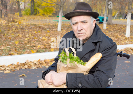 Senior man sitting in a wheelchair with a brown paper bag of groceries clutched on his lap as he waits in the road for assistance. Stock Photo