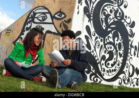 Two mixed race teenage students sitting beside some urban graffiti Stock Photo