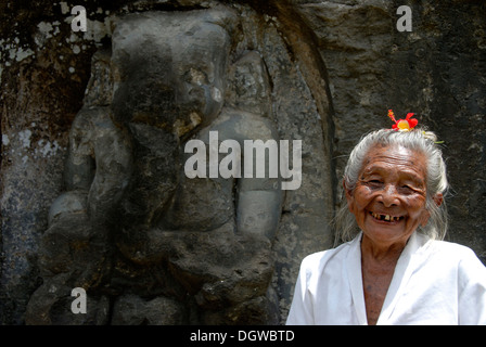 Bali Hinduism, portrait of an old Balinese woman with flower in her hear laughing in front of rock relief, God Ganesh or Ganesha Stock Photo