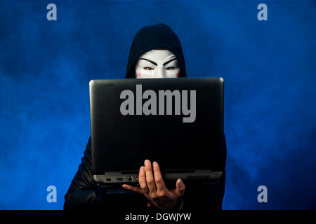 Young man wearing an anonymous mask with a hood, holding a laptop Stock Photo