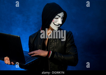 Young man wearing an anonymous mask with a hood and a black leather jacket, holding a laptop and looking to the side Stock Photo