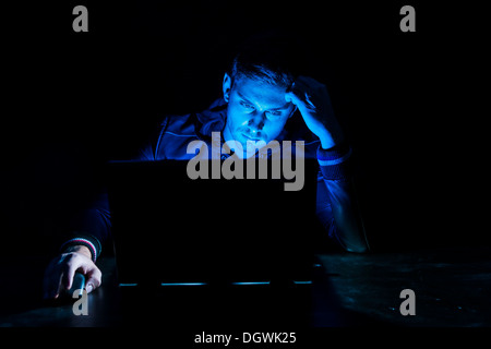 Young man sitting in front of a laptop in the dark, illuminated only by the monitor Stock Photo