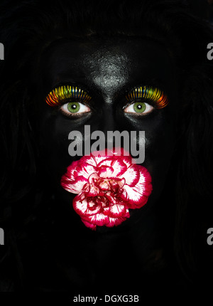 Face of a young woman painted with black facepaint, with multi-coloured eyelashes and a flower in her mouth, Austria Stock Photo