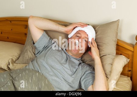 Horizontal photo of mature man holding wash cloth to his forehead along with thermometer in mouth while lying in bed Stock Photo