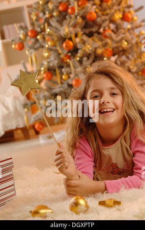 Smiling girl lying on the ground with a golden star and gifts in front of a Christmas tree Stock Photo