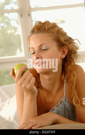 Blonde woman, 25-35 years, is propped up on her belly in bed holding an apple Stock Photo