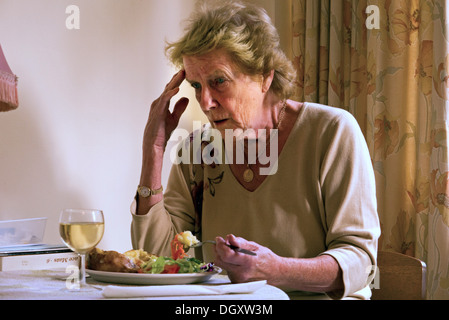 An elderly woman with Dementia and Alzheimer's Disease sitting in her home eating a meal looking confused and worried. Stock Photo