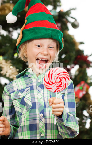 A happy laughing boy dressed as Santas helper or an elf is holding and looking at a big red lollipop. Isolated on white. Stock Photo