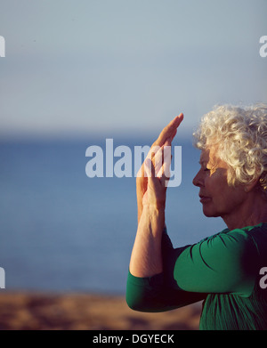 Old woman stretching her arms by the sea. Senior caucasian woman exercising on beach in morning. Fitness and healthy lifestyle Stock Photo