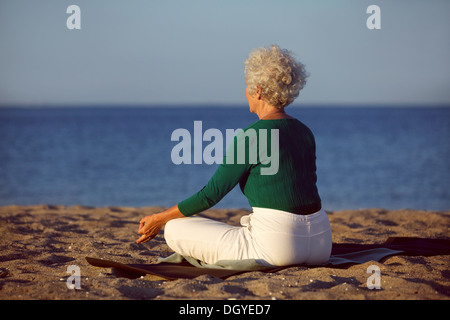 Side view of senior woman in meditation on the beach. Elderly woman sitting on the beach in lotus pose doing relaxation exercise Stock Photo