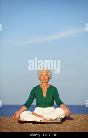Senior woman practicing morning yoga with ocean in background. Old caucasian woman doing relaxation exercise on beach. Stock Photo
