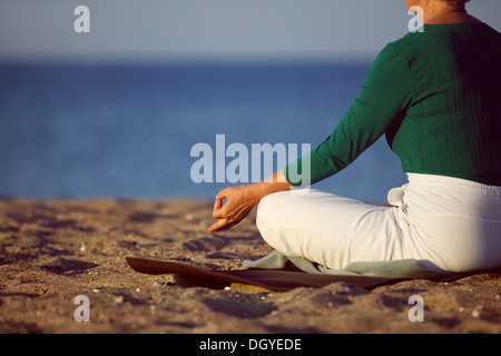 Cropped image of senior woman in meditation on sandy beach. Elderly woman sitting on the beach in lotus pose doing relaxation Stock Photo