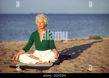 Elderly woman sitting on exercise mat doing meditation in lotus pose on the beach. Old woman doing relaxation exercise Stock Photo