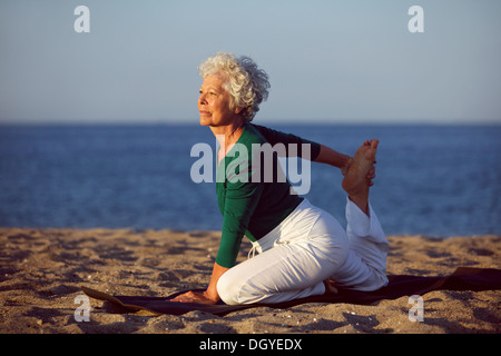 Senior woman in stretching position by the sea at morning. Elderly woman doing yoga on the beach. Stock Photo