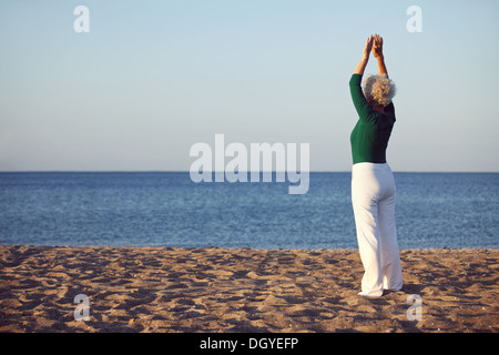 Senior woman raises her arms over her head in a stretching position while standing by the sea at morning. Old woman exercising Stock Photo