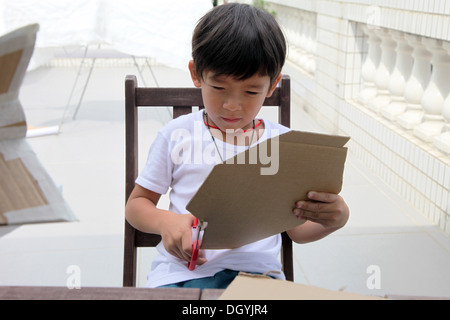 It's a photo of a little asian boy whi is doing DIY work with Cardboard to do a Rocket. He cuts, pastes and paints it. He is pro Stock Photo