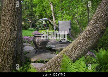 View through two tree trunks of a planter and a chair on a raised platform in a landscaped backyard garden in spring, Quebec, Stock Photo