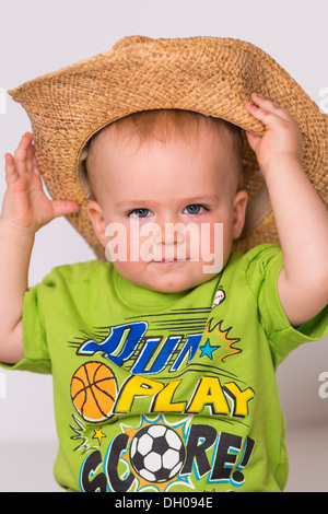 Male toddler child, 18 months old, caucasian ethnicity, is grabbing a straw cowboy hat he's wearing, which is way too large. Stock Photo
